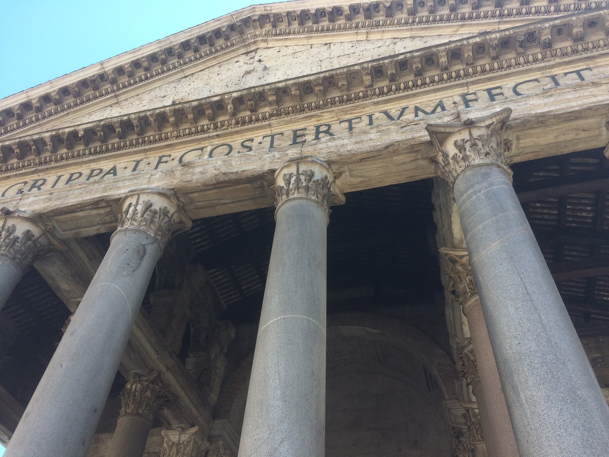 Looking up at a classical portico with granite columns and a Latin inscription ending in TERTIVM FECIT, numbered markers on four clues