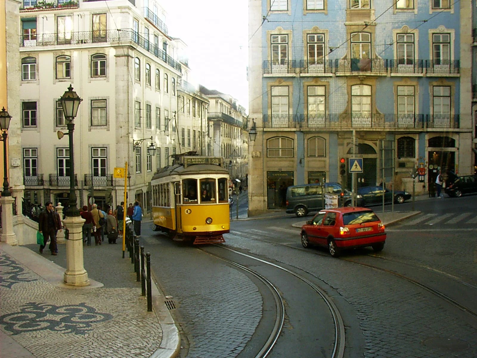 A yellow tram on curving tracks at a Lisbon intersection, with tiled facades, balconies, and patterned paving