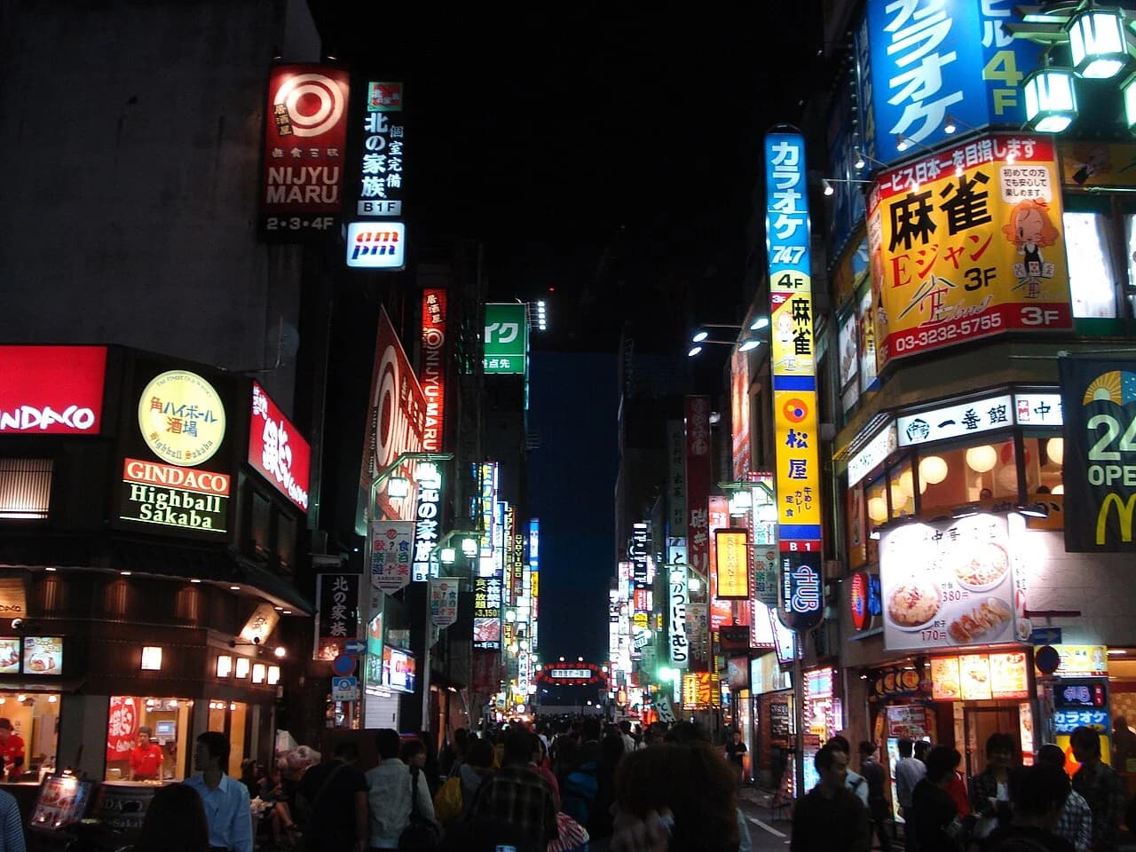 A narrow street in Shinjuku at night, with illuminated Japanese shop signs on both sides and pedestrians below